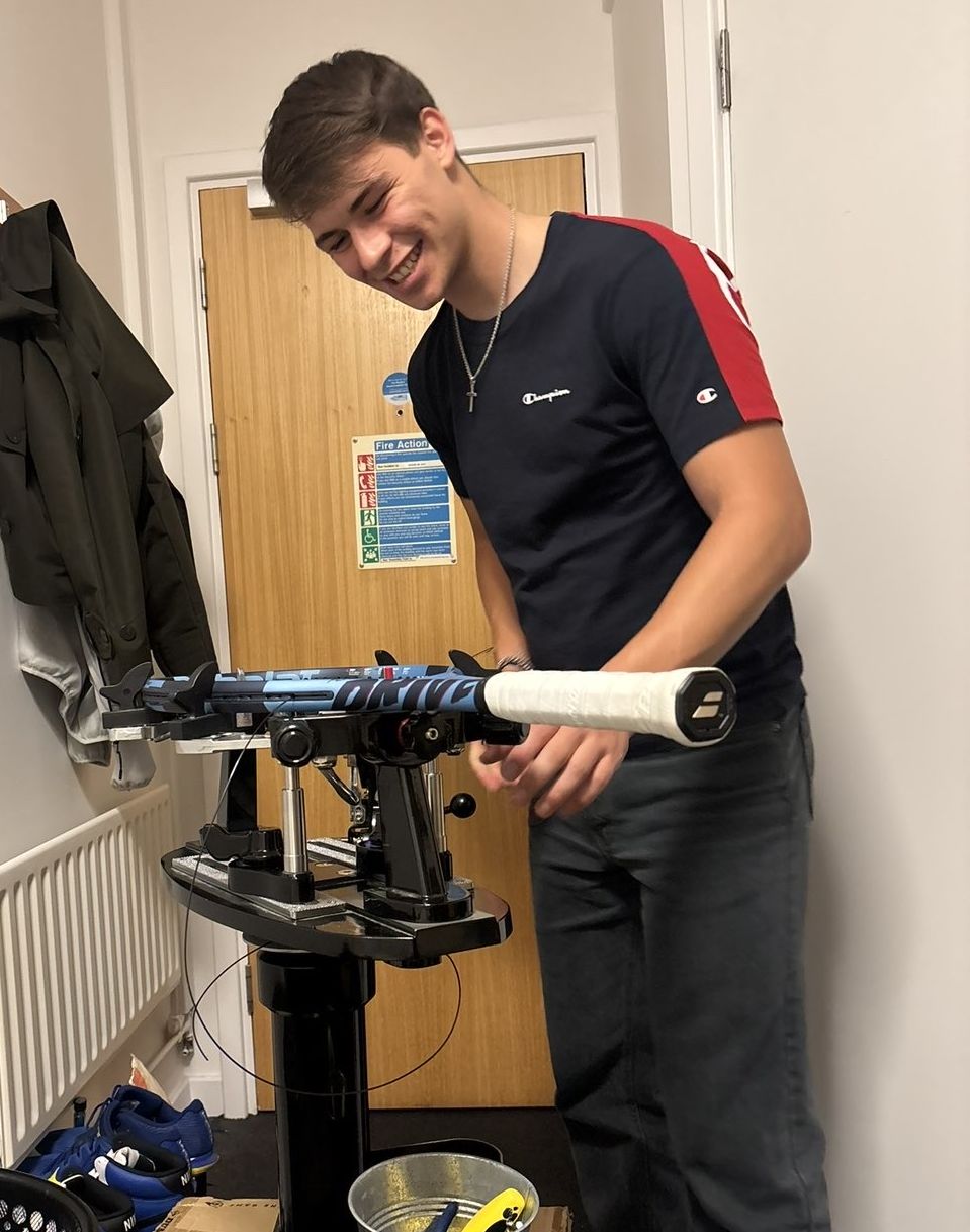 Luka Matellini laughing while stringing a Babolat tennis racket